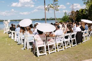 Outdoor wedding ceremony with guests seated in white chairs holding parasols by a lakeside on a sunny day.