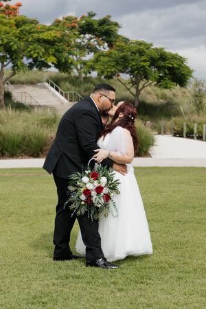 Bride and groom share a kiss on green lawn, she holds red and white bouquet, he wears black suit, trees in background.