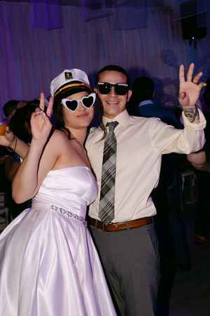 Bride and groom dancing at wedding reception wearing fun sunglasses and captain hat, posing playfully on the dance floor.