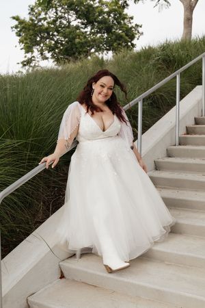 Plus-size bride in white tulle wedding gown with lace bodice descending outdoor stone stairs surrounded by greenery.