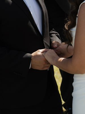 Bride and groom holding hands during outdoor wedding ceremony, close-up of couple in formal attire exchanging rings.