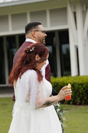 Bride in white lace gown with veil and groom in burgundy suit stand together outdoors at wedding venue.
