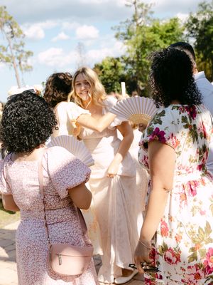 Bride in white dress holding fan smiles at guests in floral dresses at outdoor wedding celebration.