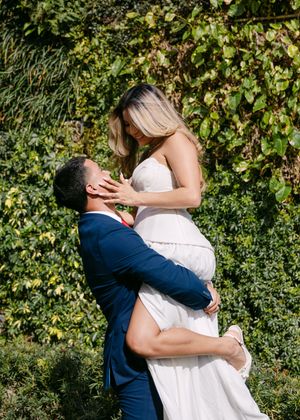 Groom in navy suit lifts bride in white wedding gown for a romantic kiss outdoors against lush green ivy.