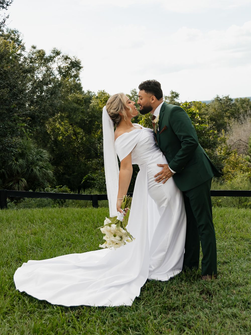 Bride and groom share a romantic kiss outdoors, she in a white gown with veil, he in a dark green suit.