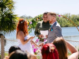 Couple exchanges vows outdoors at waterfront wedding ceremony with floral arch, officiant, and guests on sunny day.