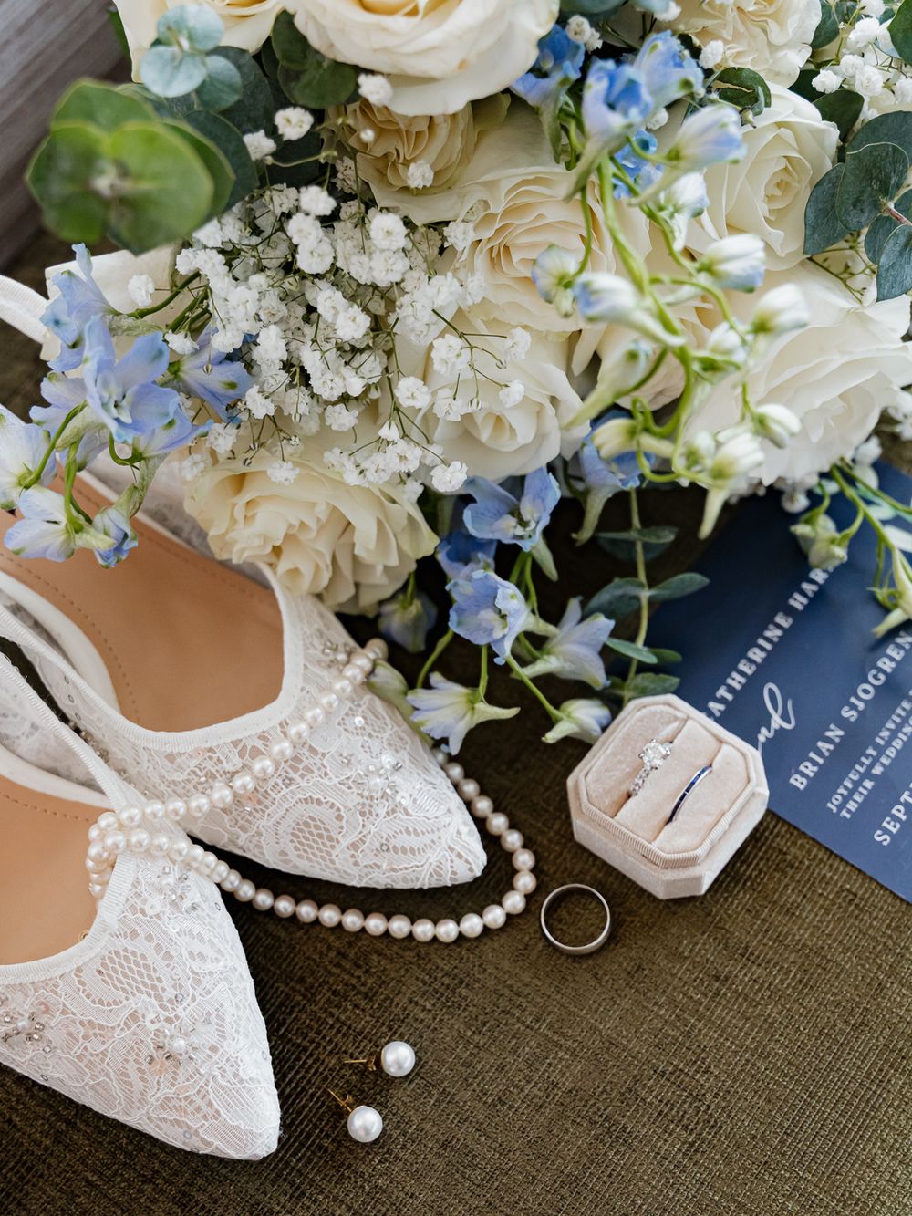 Elegant wedding flat lay with lace bridal shoes, pearl necklace, engagement ring, earrings, floral bouquet, and navy invitation.