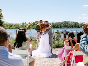 Bride and groom share first kiss at outdoor waterfront wedding ceremony with guests watching on a sunny day.