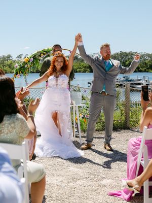 Joyful newlyweds celebrate after outdoor waterfront wedding ceremony, bride in white gown with floral crown, groom in gray suit.