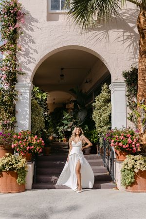 Woman in white dress posing under floral archway with lush greenery and pink flowers at elegant building entrance.