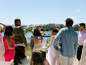 Guests celebrate at an outdoor waterfront wedding ceremony on a sunny day, with people hugging and cheering.
