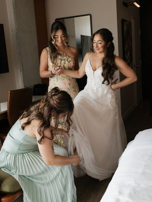 Bride in white lace gown being helped by two bridesmaids in floral and mint dresses in a hotel room.