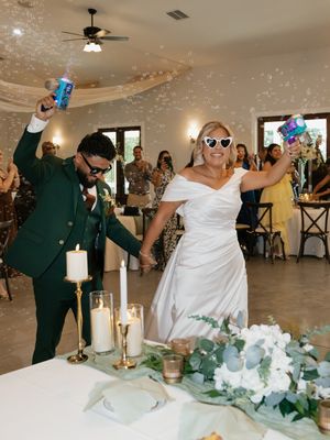 Joyful newlyweds celebrate at wedding reception, groom in green suit and bride in white gown holding sparklers near cake table.