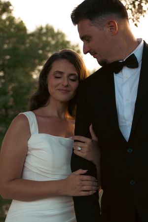 Bride in white gown and groom in black tuxedo share a tender moment outdoors at golden hour.