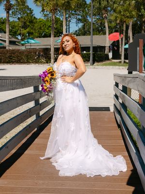 Beautiful bride in white strapless wedding gown holding colorful bouquet on sunny beach boardwalk with palm trees.