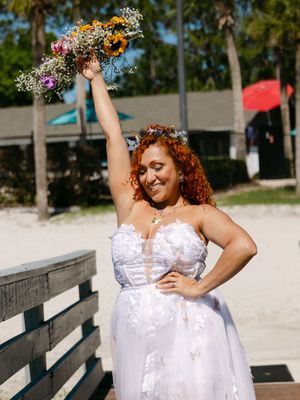 Joyful bride in white lace gown raises colorful wildflower bouquet on beach boardwalk, wearing floral crown.