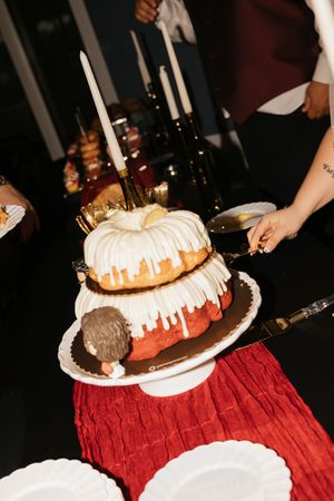 Two-tier bundt cake with white icing drizzle, candles, and figurine topper being cut at a celebration event.