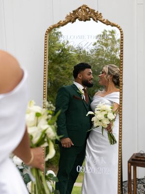 Bride and groom reflected in ornate gold mirror at outdoor wedding, bride in white gown holding bouquet, groom in dark green suit.