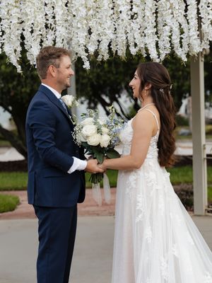 Bride and groom holding hands under white floral arch, smiling at each other during outdoor wedding ceremony.