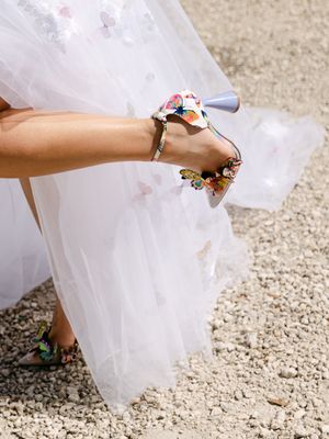 Elegant bridal heels with colorful floral and butterfly embellishments paired with a flowing white tulle wedding gown outdoors.