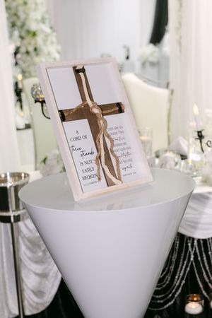Framed Christian cross with Bible verse on white pedestal at elegant wedding reception decor.