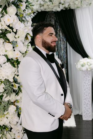 Groom in white tuxedo jacket with black lapels stands at floral wedding altar with white roses.