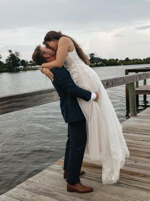 A groom lifts his bride in a white lace gown on a wooden dock waterfront wedding photo.