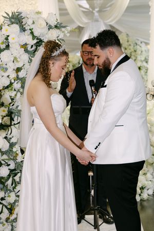 Bride and groom holding hands at floral altar during wedding ceremony with officiant in background.