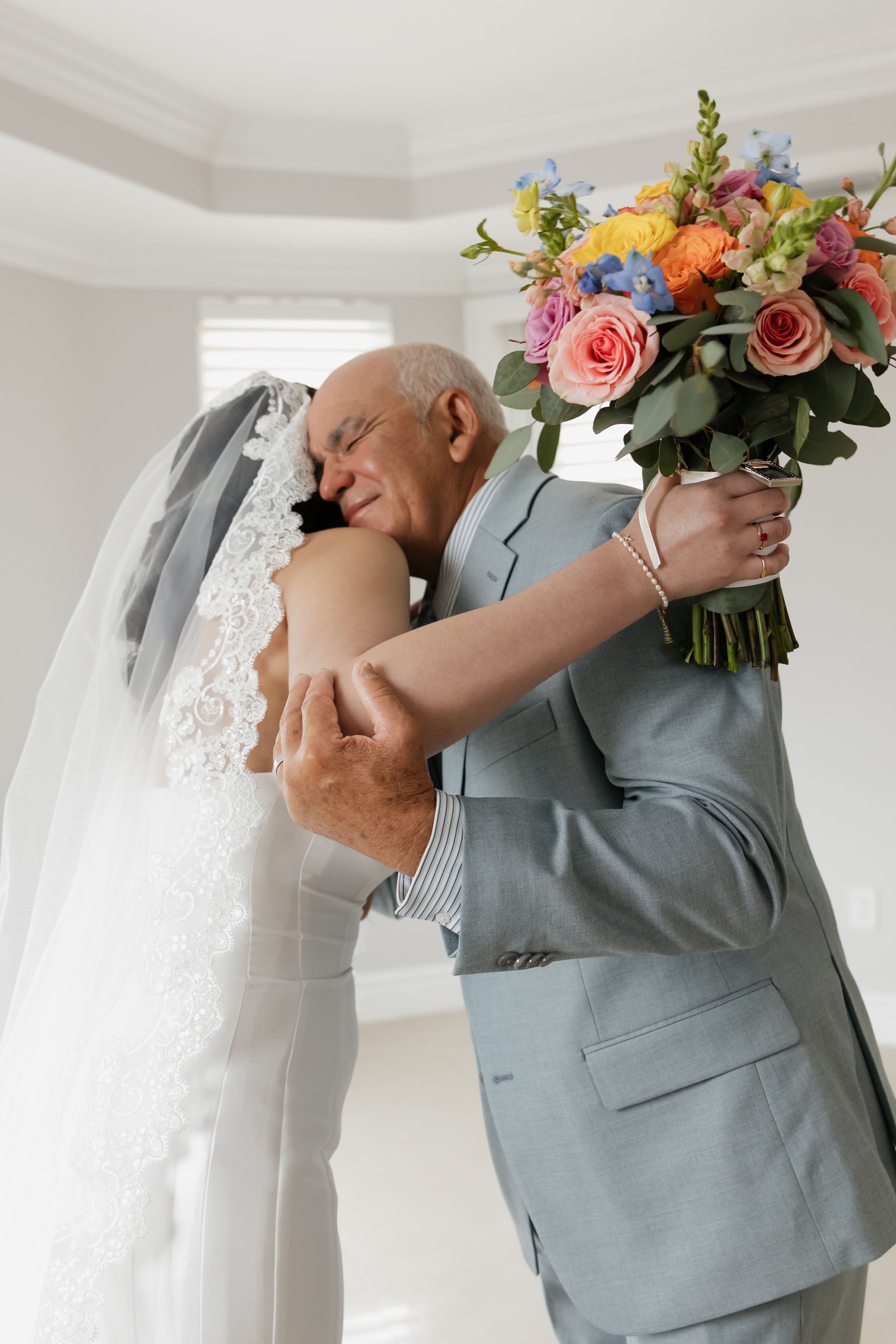 Bride and older man in gray suit share emotional hug, bride holding colorful bouquet of roses and mixed flowers.