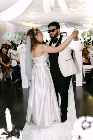 Bride and groom share first dance at elegant white wedding reception, groom wearing sunglasses and white tuxedo jacket.