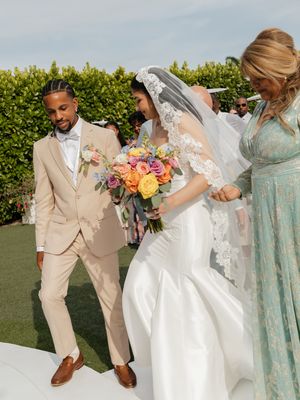 Bride in lace wedding gown holding colorful bouquet walks with groom in tan suit at outdoor ceremony.