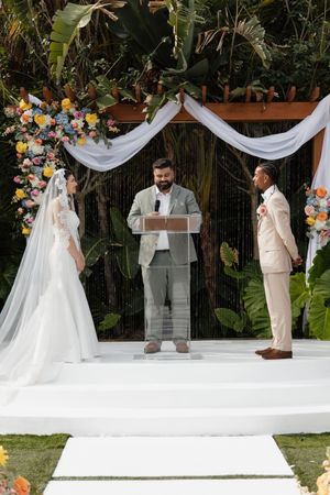 Outdoor wedding ceremony with bride in white gown, groom in beige suit, and officiant at podium under floral arch.