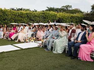Guests seated in white chairs at an outdoor wedding ceremony on a green lawn surrounded by lush hedges.