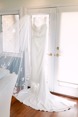 Elegant white strapless wedding gown with cathedral train and floral veil hanging on a door in a bright room.