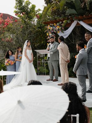 Outdoor wedding ceremony with bride in white gown, groom in beige suit, officiant, and guests holding white parasols.