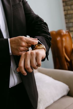 Man in black suit adjusting gold wristwatch, wearing white dress shirt, seated near sofa in elegant interior.