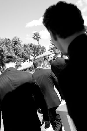 Black and white photo of men in dark suits walking toward a building with palm trees under a bright sky.