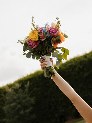 A hand raises a colorful bridal bouquet with pink, yellow, and orange roses against a green hedge backdrop.