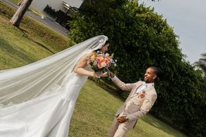 Bride in white gown with flowing veil holds colorful bouquet as groom in beige suit reaches for her hand outdoors.