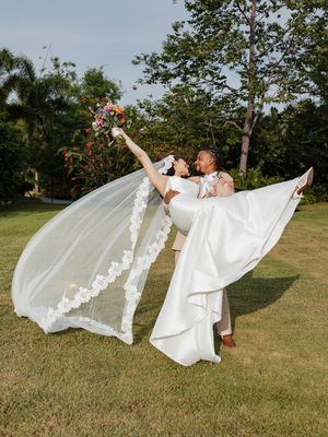 Groom lifts bride in white gown and veil, holding colorful bouquet aloft on green lawn with tropical trees.