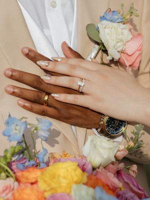 Two hands with wedding rings resting on colorful bouquet, one wearing diamond engagement ring and gold band.
