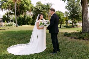 Bride in white gown with veil holds bouquet as groom in black tux reads vows outdoors on lush green lawn.