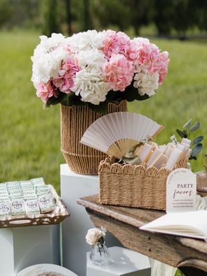 Outdoor wedding favor table with pink and white hydrangeas, wicker baskets, paper fans, and party favor sign on white pedestals.