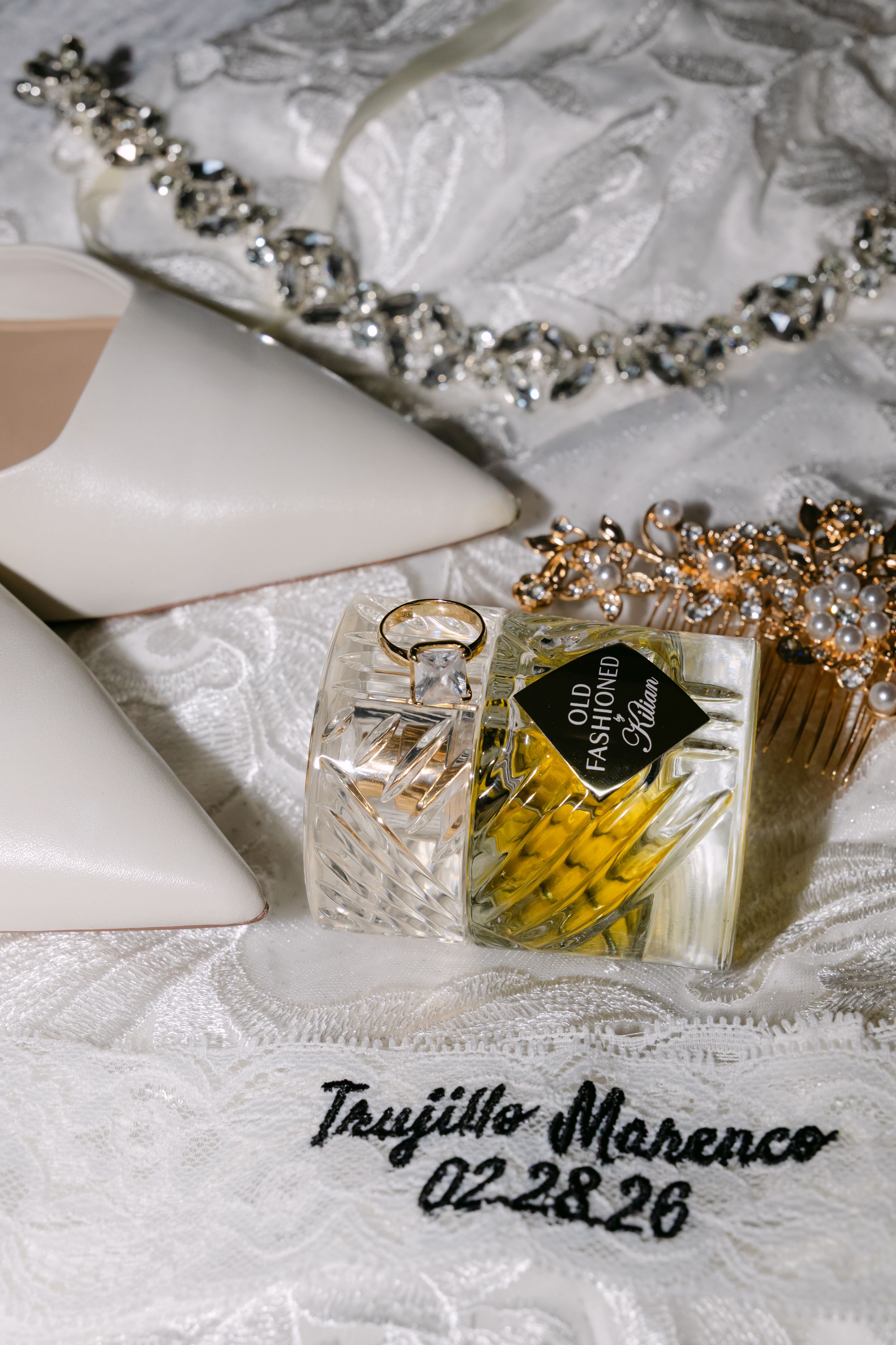 Wedding flat lay featuring bridal shoes, diamond jewelry, gold ring, perfume bottle, and embroidered handkerchief dated 02-28-26.