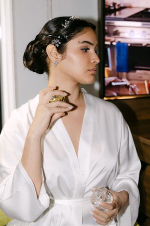 Bride in white silk robe applying perfume, wearing crystal hair pins and drop earrings while getting ready.