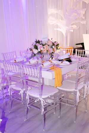 Elegant wedding reception table with crystal chairs, floral centerpiece, and soft purple lighting.