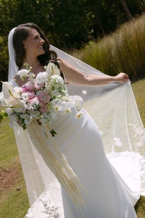 Radiant bride in white gown holding pink floral bouquet, veil flowing in breeze outdoors on sunny wedding day.