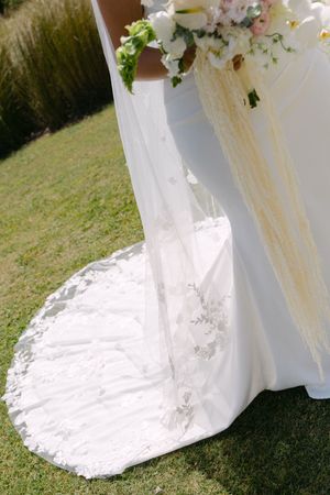 Bride in white wedding gown with lace train holding floral bouquet on green lawn.