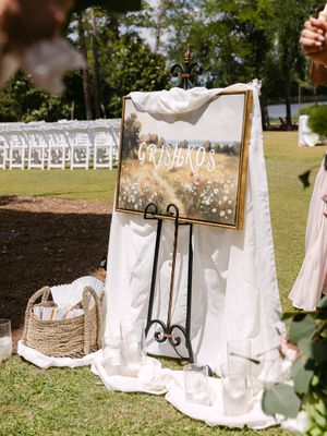 Outdoor wedding ceremony setup with floral art sign on easel reading Grishnos, white draping, and rows of chairs on green lawn.