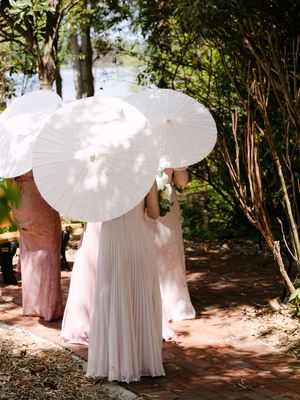 Bridesmaids in pink dresses holding white paper parasols outdoors at a garden wedding ceremony.